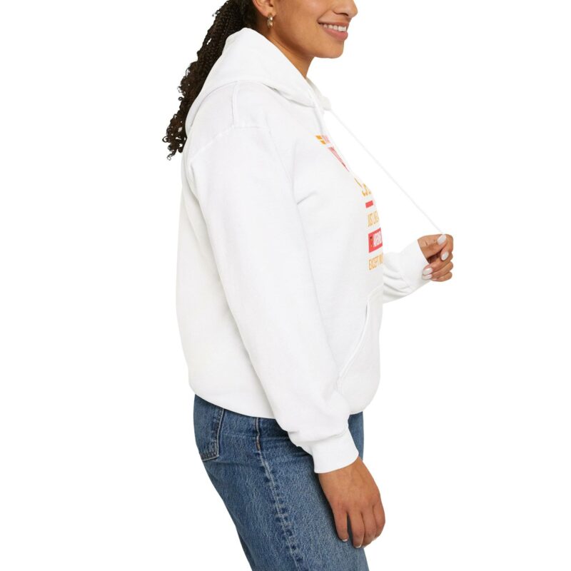 A person wearing the "I'm an Airbnb Superhost" Unisex Heavy Blend Hooded Sweatshirt with multicolored text on the front and blue jeans, viewed from the side. The person is pulling one of the hoodie strings with their right hand.