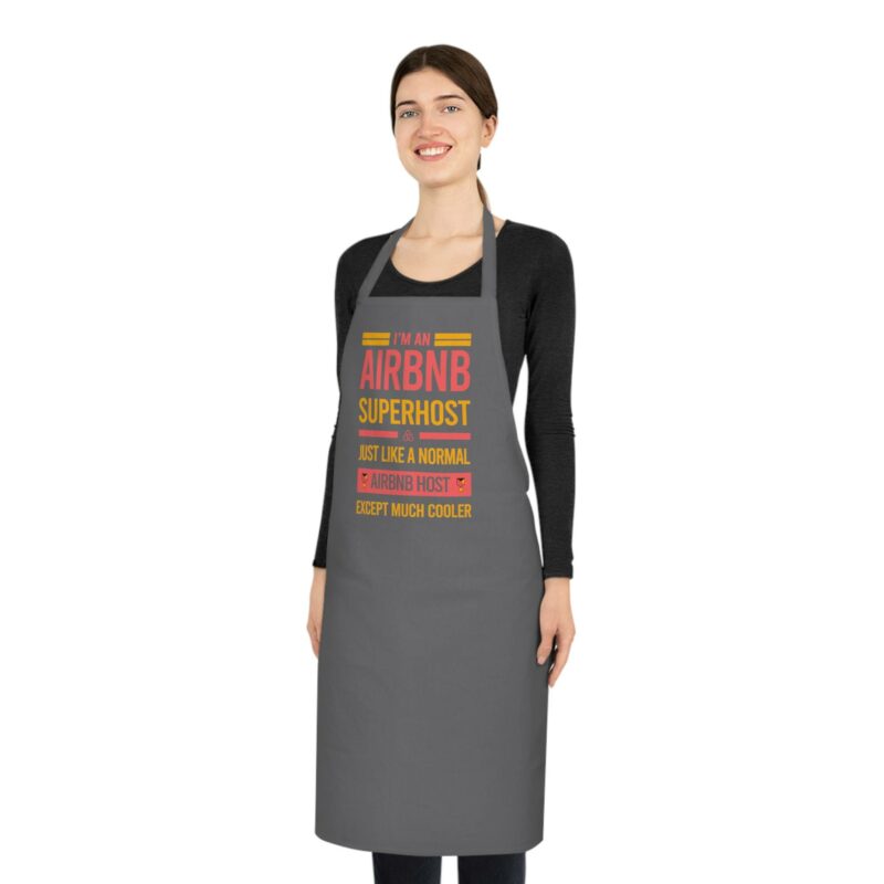 A person standing and smiling wears the "I'm an Airbnb Superhost Cotton Apron" in grey, proudly displaying the text "I'm an Airbnb Superhost, just like a normal Airbnb host except much cooler.