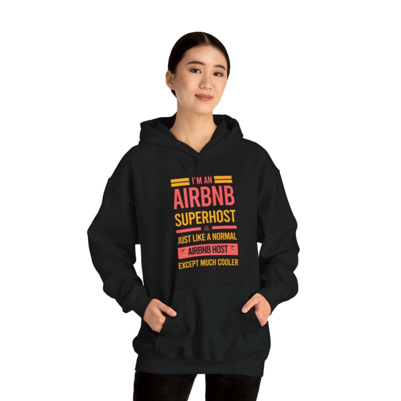 A person wearing the "I'm an Airbnb Superhost Unisex Heavy Blend Hooded Sweatshirt," featuring a colorful text that reads "I'm an Airbnb Superhost. Just like a normal Airbnb host except much cooler," standing and smiling.