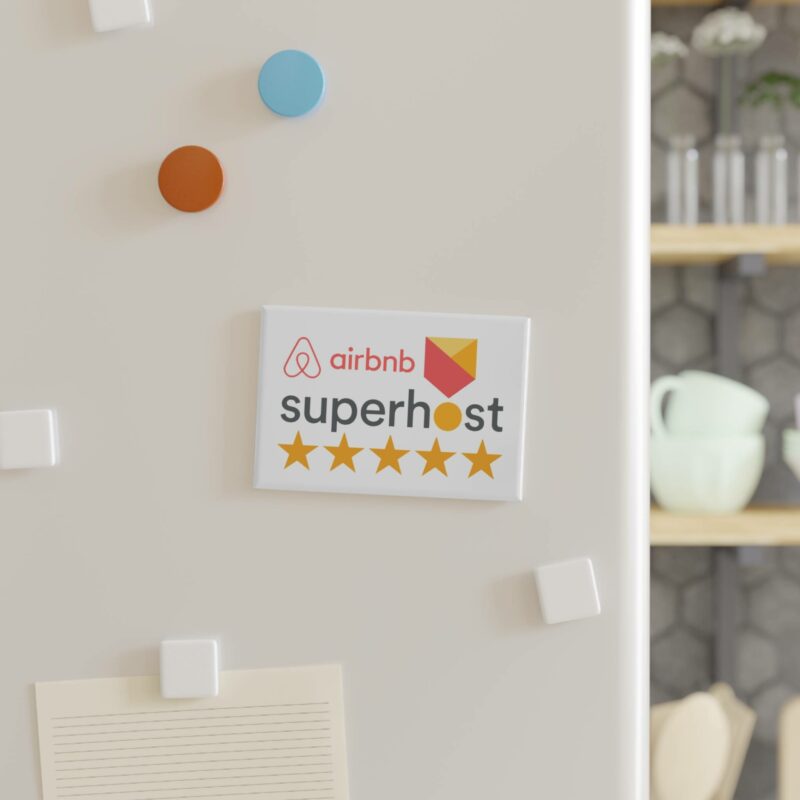 A refrigerator door featuring an "I'm an Airbnb Superhost" rectangular button magnet showcasing five stars, surrounded by vibrant round and square white magnets. Kitchen items are visible in the blurry background.