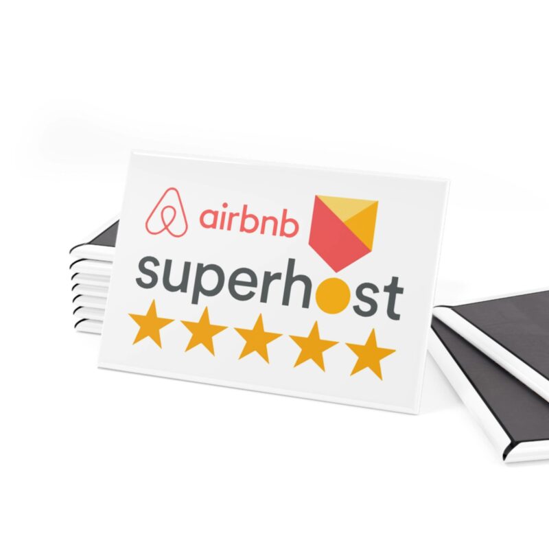 A white "I'm an Airbnb Superhost" button magnet, featuring the Airbnb logo and a five-star rating, rests atop stacked documents. Two black notebooks are visible to the right.