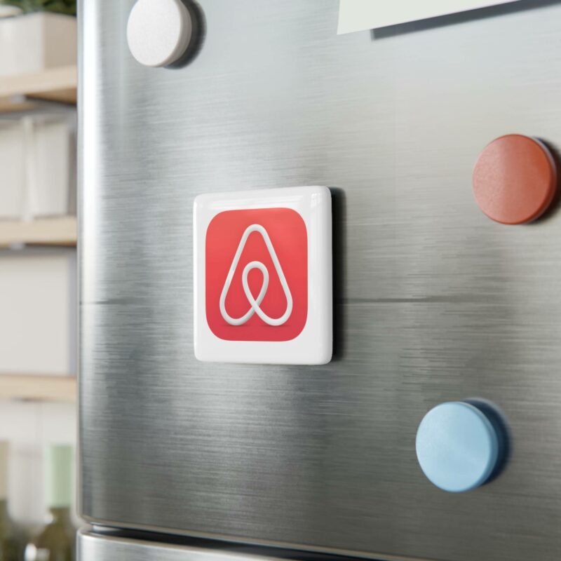 A red and white logo, resembling the Airbnb logo, is displayed on a metallic surface with three colorful magnets (white, red, and blue) nearby. Shelves with items are visible in the background. The red magnet is an Airbnb Porcelain Magnet, Square.