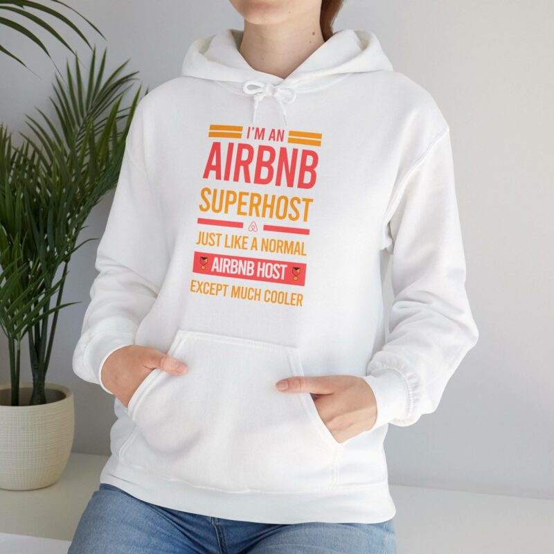 An individual is wearing an "I'm an Airbnb Superhost" Unisex Heavy Blend Hooded Sweatshirt, which features the text, "Just like a normal Airbnb host except much cooler." A potted plant can be seen in the background.