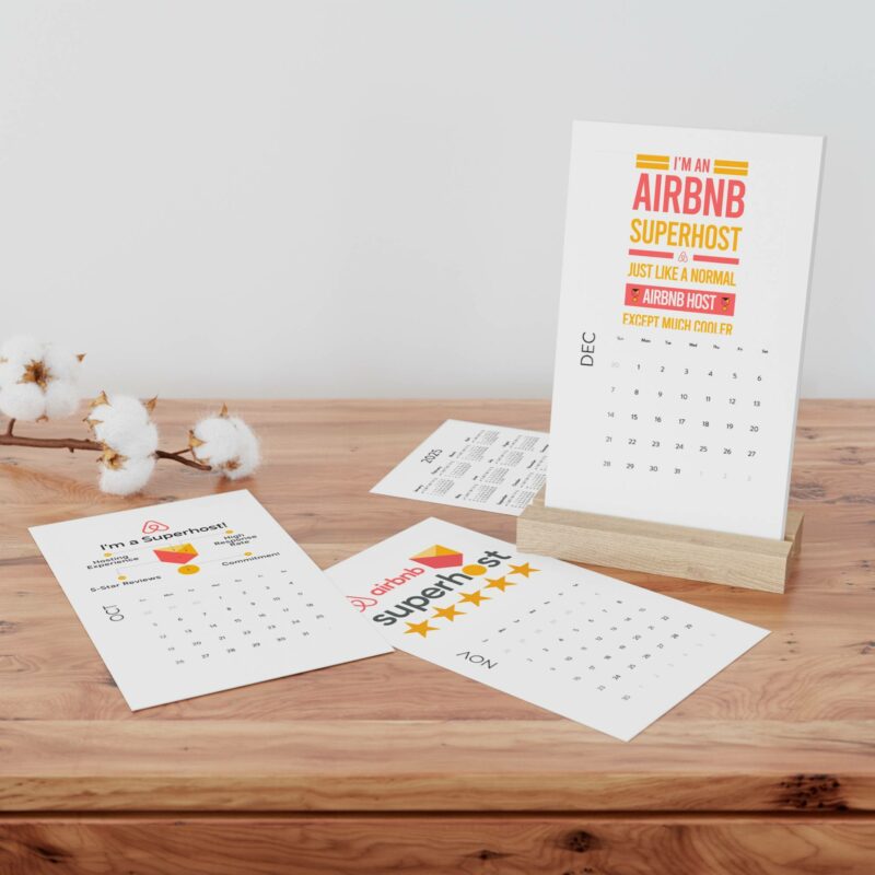 Displayed on a wooden stand, the Vertical Desk Calendar (2022) showcases its December card, which features the message "I'M AN AIRBNB SUPERHOST" in bold yellow and orange text. Additional monthly cards are spread out on the wooden table, complemented by cotton buds that adorn the background.