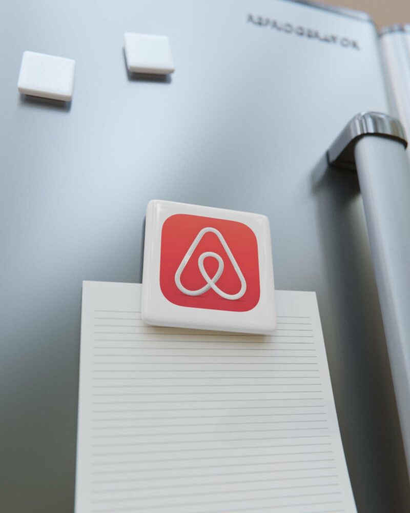 A rectangular magnet with a red and white icon is holding a lined paper on a metallic refrigerator door, which also has three Airbnb Porcelain Magnets, Square.