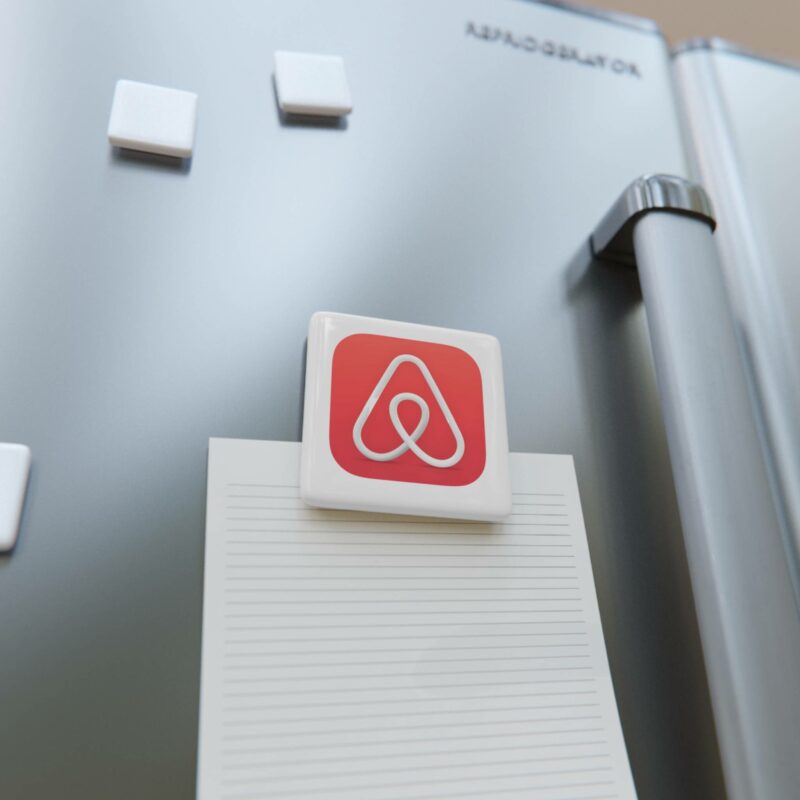 A rectangular magnet with a red and white icon is holding a lined paper on a metallic refrigerator door, which also has three Airbnb Porcelain Magnets, Square.