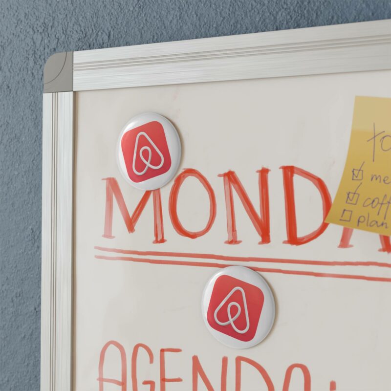 Whiteboard with the word "Monday" written in red and two Airbnb Button Magnets featuring a round logo. A sticky note with a checklist is partially visible in the top right corner.