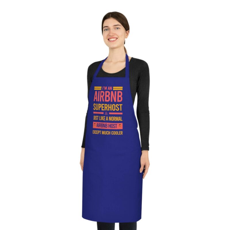 Person smiling while wearing the "I'm an Airbnb Superhost Cotton Apron" featuring the text "I’m an Airbnb Superhost, just like a normal Airbnb host except much cooler," over a black long-sleeve shirt.