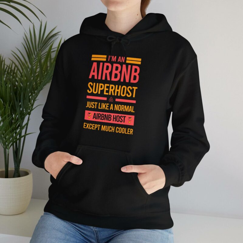 A person wearing the "I'm an Airbnb Superhost Unisex Heavy Blend Hooded Sweatshirt," featuring the slogan, "I'm an Airbnb Superhost. Just like a normal Airbnb host except much cooler," stands near a plant.