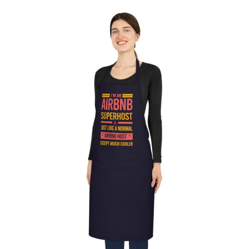 A woman stands smiling, wearing an "I'm an Airbnb Superhost Cotton Apron," which features the text "I'M AN AIRBNB SUPERHOST... JUST LIKE A NORMAL AIRBNB HOST EXCEPT MUCH COOLER." She has dark hair tied back and wears a black long-sleeve shirt.