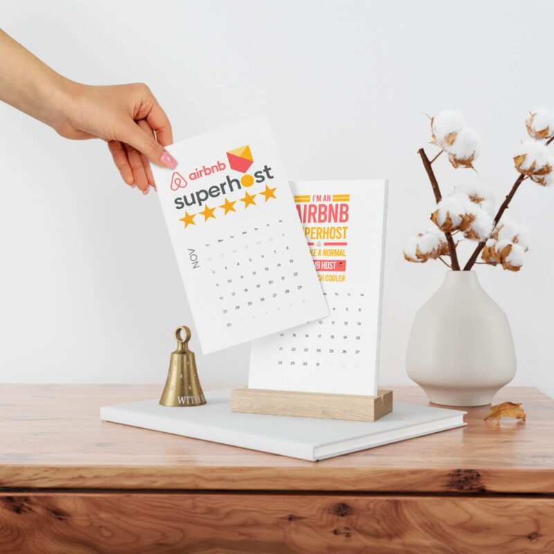 A hand holding a calendar page marked "Airbnb Superhost" with five stars. The scene includes a desk with a potted cotton plant, a bell, and the Vertical Desk Calendar (2022) on a wooden stand.