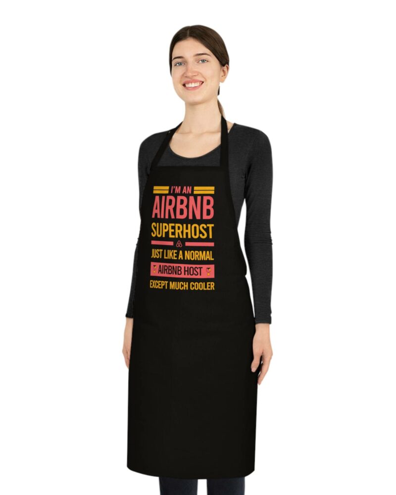 A person smiles while standing against a plain white background, wearing the "I'm an Airbnb Superhost Cotton Apron," which features the text, "I'm an Airbnb superhost. Just like a normal Airbnb host, except much cooler.