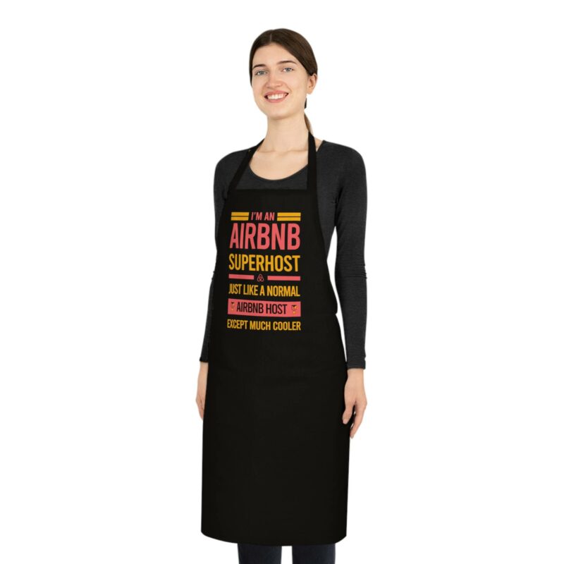 A person smiles while standing against a plain white background, wearing the "I'm an Airbnb Superhost Cotton Apron," which features the text, "I'm an Airbnb superhost. Just like a normal Airbnb host, except much cooler.