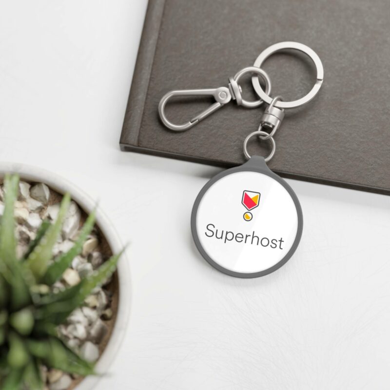 The "I'm an Airbnb Superhost Keyring Tag" rests on a table, accompanied by a potted plant and a closed notebook, making it an ideal addition for any Airbnb hosting enthusiast.