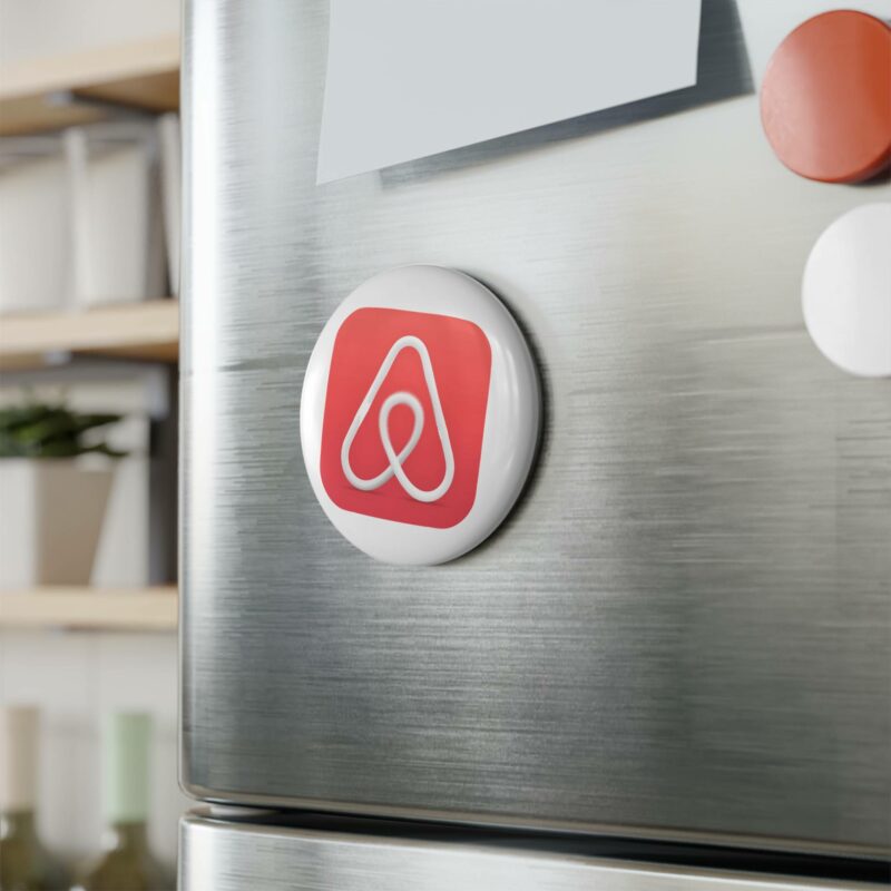 An Airbnb Button Magnet, Round, is on a stainless steel refrigerator door, next to other magnets and papers.