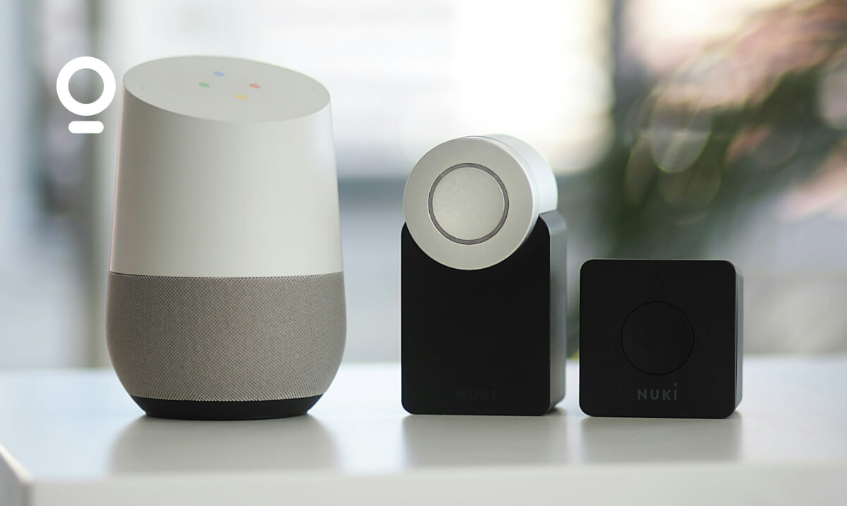 A Google Home smart speaker is placed next to two Nuki devices on a white surface.