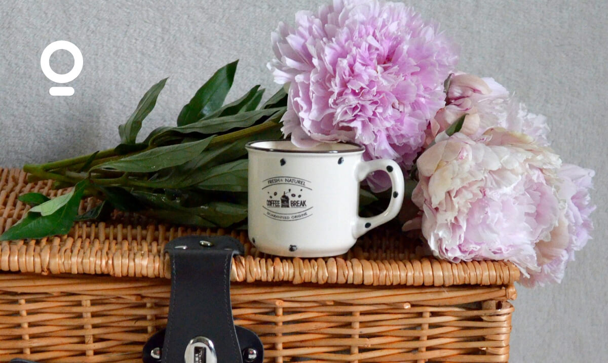 A white mug and pink peonies rest on a wicker basket with a leather strap and latch.