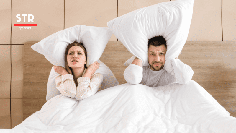 Airbnb Hosting A man and woman in bed with pillows covering their heads, utilizing NoiseAware to monitor their Airbnb party squasher.
