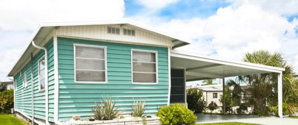 A small turquoise house with a white trim and carport showcases how selecting the perfect manufactured home floor plans for your family can create cozy spaces. Two windows on the side and plants in front add charm, while the partly cloudy sky enhances the inviting atmosphere.