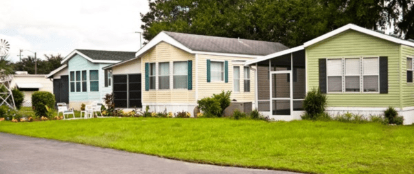 A row of colorful mobile homes with green lawns and gardens sits gracefully under a partly cloudy sky, each offering unique features tailored for selecting the perfect manufactured home floor plans for your family.