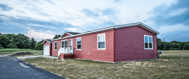 A red, single-story house with white trim and a small porch, surrounded by a grassy area and trees in the background. Under the cloudy sky, it stands as an ideal example of selecting the perfect manufactured home floor plans for your family.
