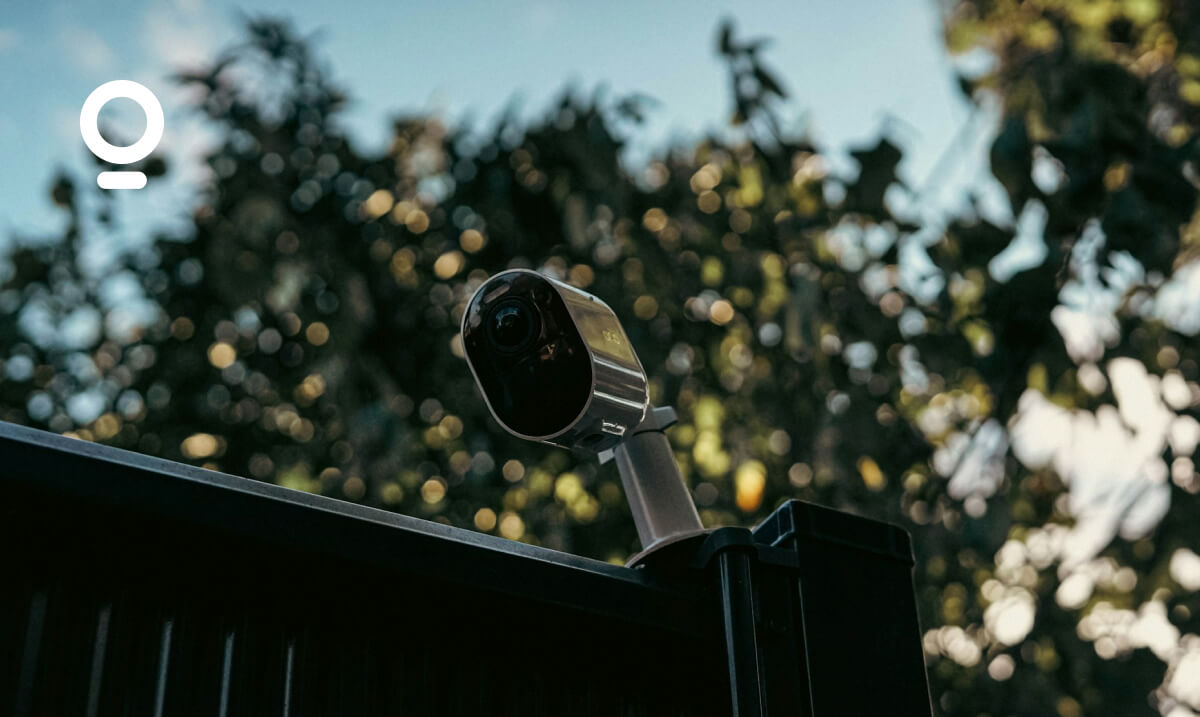 A security camera mounted on a black fence with trees in the background is shown in this image. The logo in the top left corner is of the company Olist.