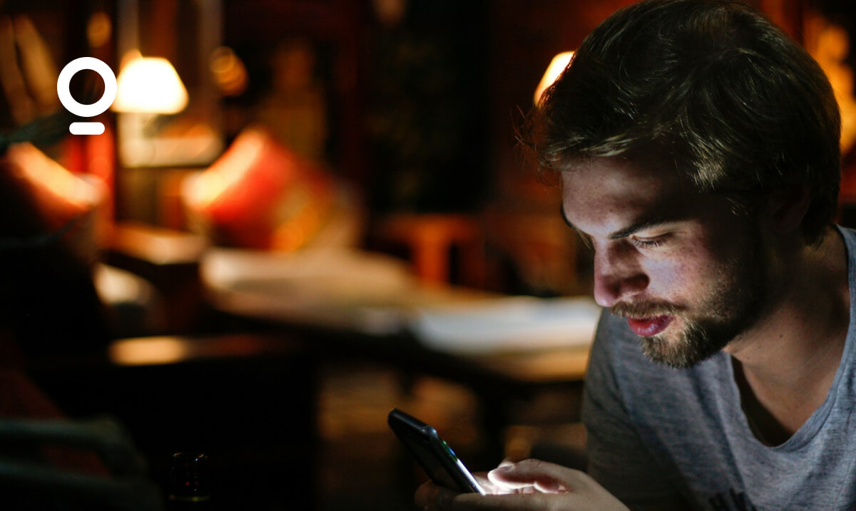 A man with a beard and casual clothes looks at his smartphone in a dimly lit room with a sofa and lamps in the background.