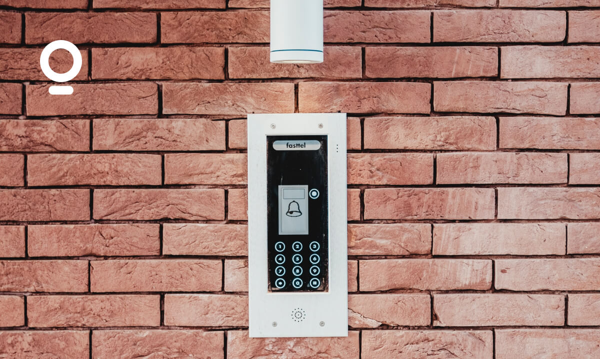 A wall-mounted intercom system with keypad on a red brick wall, featuring a small display and a doorbell button. A white cylindrical fixture is installed above the intercom.