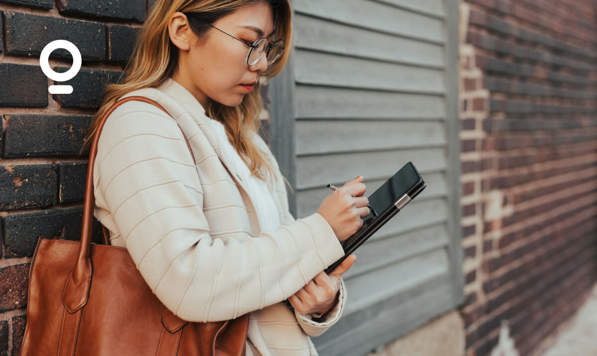Person with glasses uses a tablet while leaning against a brick wall. They have a brown bag over their shoulder and are wearing a light-colored jacket. A circular logo is visible on the wall.