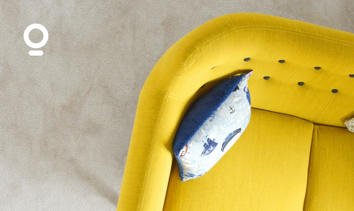 Top view of a yellow sofa with rounded edges and a blue, patterned throw pillow. A white circular logo is present in the top left corner of the image.