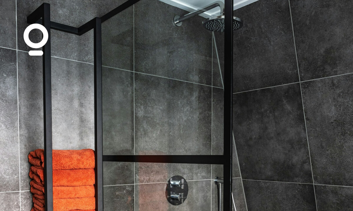 Modern bathroom with dark tiled walls, a glass-enclosed shower with a rainfall showerhead, and a stack of orange towels placed on a wall-mounted shelf.