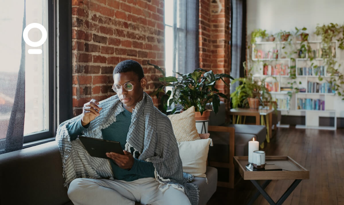 A person sits on a couch in a room with exposed brick walls, reading a tablet and holding a pen, surrounded by plants and candles.