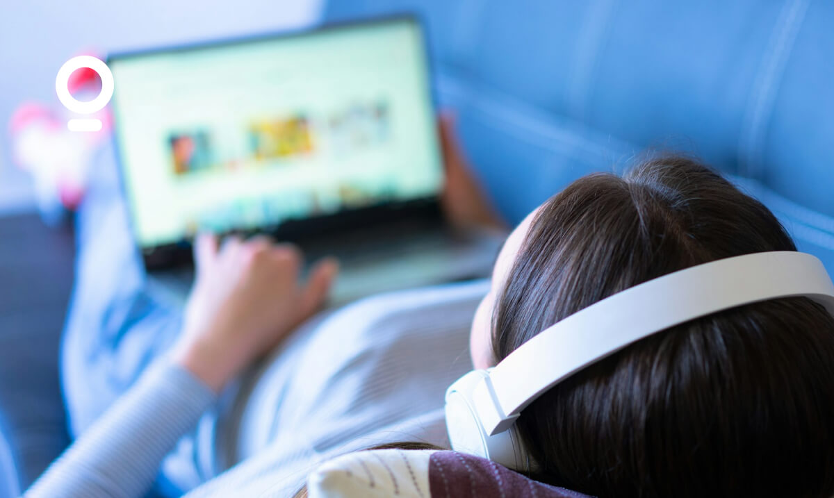 Person wearing white headphones and lying on a blue sofa, using a laptop.